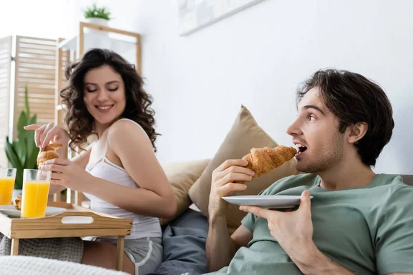 Чоловік їсть круасан біля щасливої жінки під час сніданку в ліжку — стокове фото