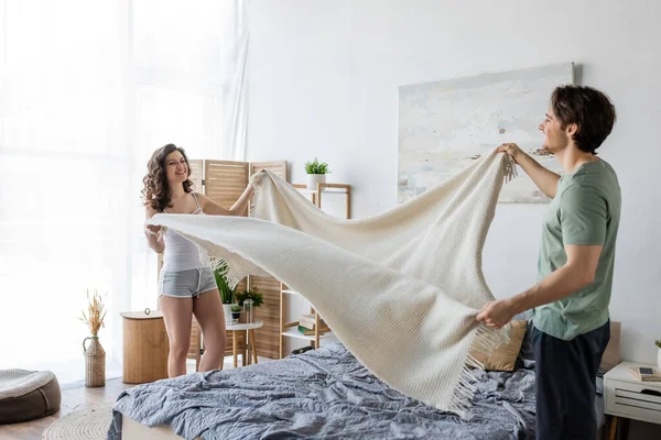 Feliz jovem casal cobrindo cama com cobertor — Fotografia de Stock