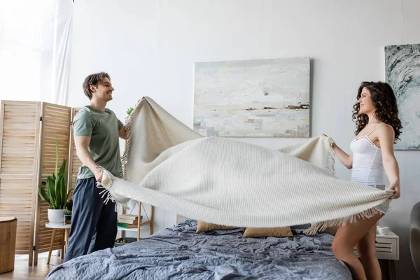 Side view of smiling young couple covering bed with blanket — Stock Photo