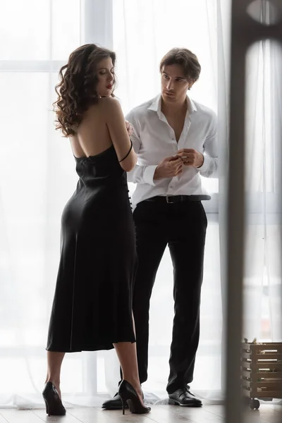 Чоловік розстібає білу сорочку біля спокусливої жінки в чорній сукні ковзання — стокове фото