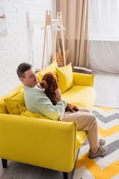 Улыбающийся подросток с синдромом Дауна держит пуделя на диване дома — стоковое фото