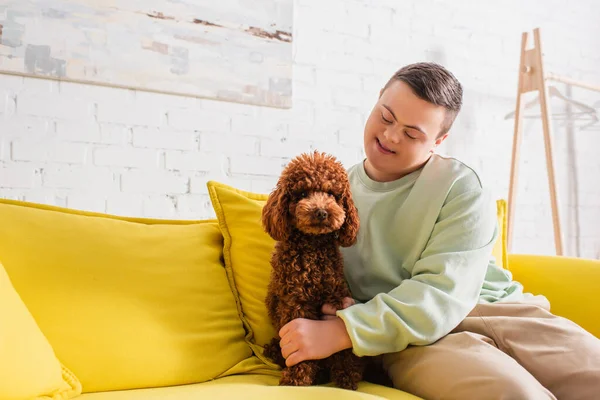 Adolescent joyeux avec le syndrome du duvet caressant caniche sur le canapé à la maison — Photo de stock