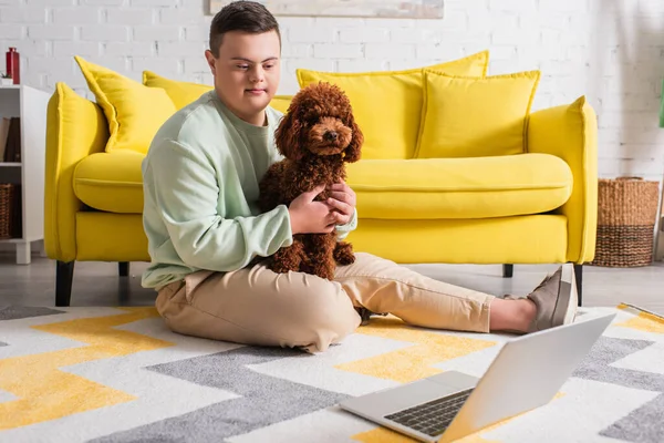 Adolescent avec le syndrome du duvet caressant caniche près de l'ordinateur portable sur le sol à la maison — Photo de stock