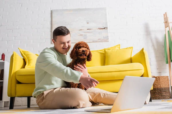 Smiling teenager with down syndrome pointing at laptop near poodle at home — Stock Photo