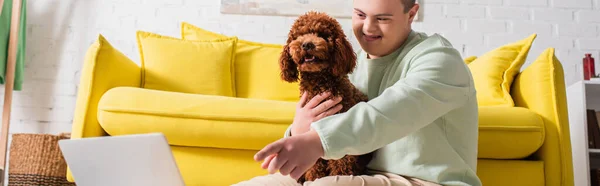 Positiver Teenager mit Down-Syndrom, der mit dem Finger auf Laptop zeigt und Pudel zu Hause umarmt, Banner — Stockfoto