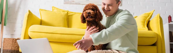 Adolescente positivo con sindrome di Down che punta con il dito al computer portatile e abbraccia barboncino a casa, banner — Foto stock