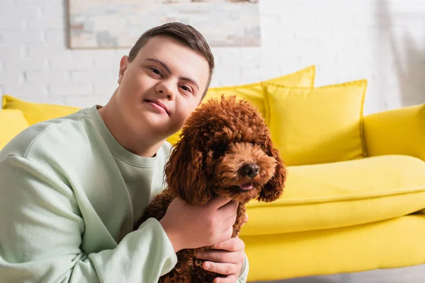 Adolescente chico con síndrome de Down abrazando caniche y mirando a la cámara en casa - foto de stock