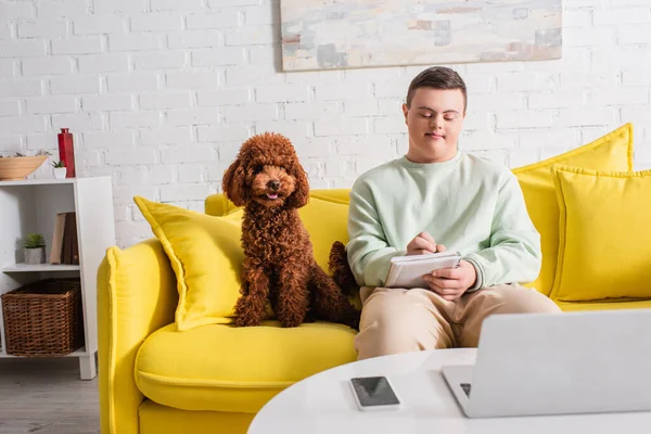 Teenager mit Down-Syndrom schreibt auf Notizbuch neben Gerät und Pudel zu Hause — Stockfoto