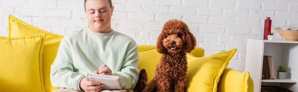 Teen boy with down syndrome writing on notebook near poodle on couch, banner — Stock Photo