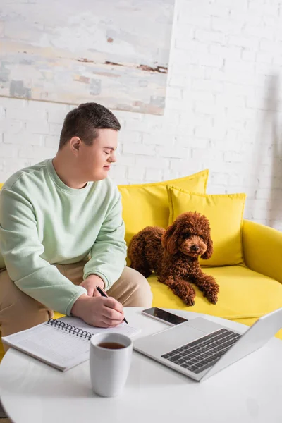 Visão lateral de adolescente sorridente com síndrome de down escrevendo no caderno e olhando para poodle perto de gadgets em casa — Fotografia de Stock