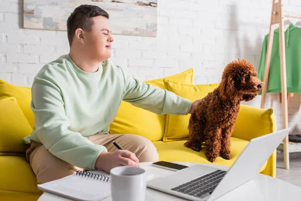 Adolescente com síndrome de down escrevendo em caderno e poodle de estimação perto de gadgets em casa — Fotografia de Stock