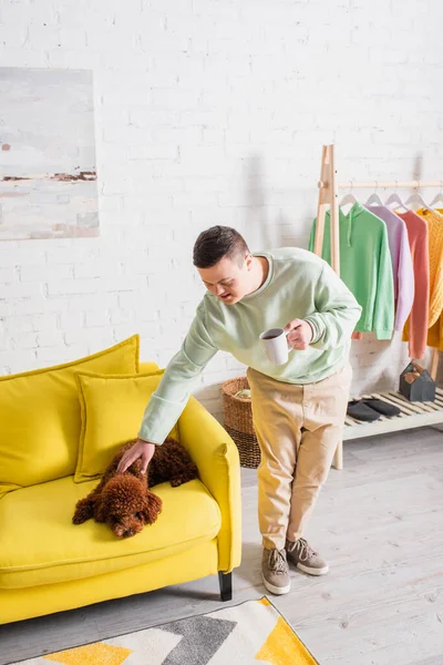 Teenager with down syndrome holding cup and petting poodle on couch at home — Stock Photo