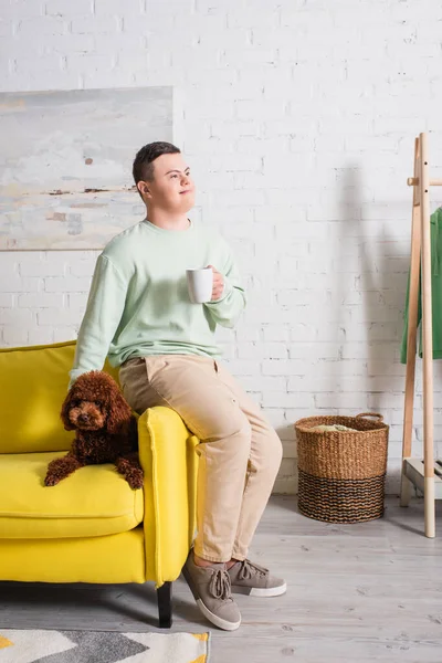 Garçon souriant avec syndrome duvet tenant une tasse près du caniche sur le canapé à la maison — Photo de stock