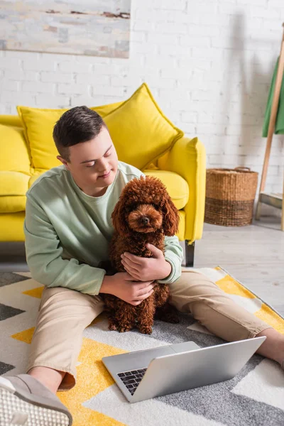 Adolescent avec le syndrome du duvet étreignant caniche près d'un ordinateur portable sur le sol à la maison — Photo de stock