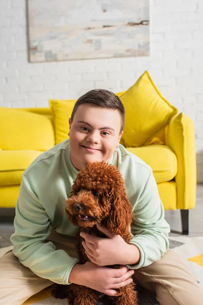 Sonriente adolescente con síndrome de Down abrazando caniche y mirando a la cámara en casa - foto de stock