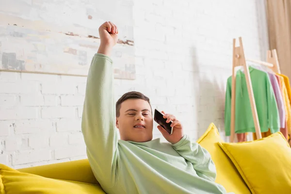 Adolescente chico con síndrome de Down sosteniendo teléfono inteligente y mirando a la cámara en el sofá en casa - foto de stock