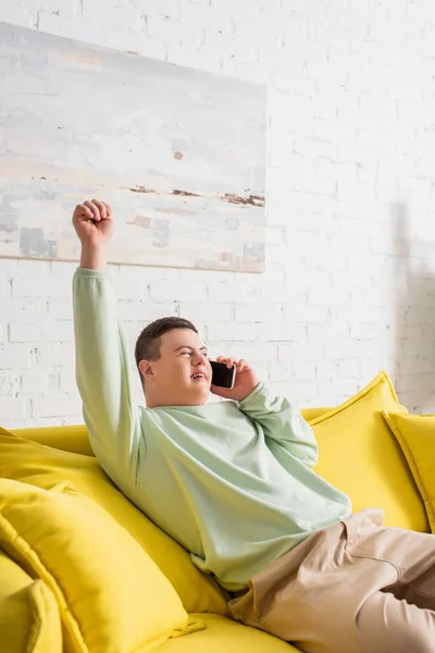 Emocionado adolescente con síndrome de Down hablando en el teléfono inteligente en el sofá en casa - foto de stock