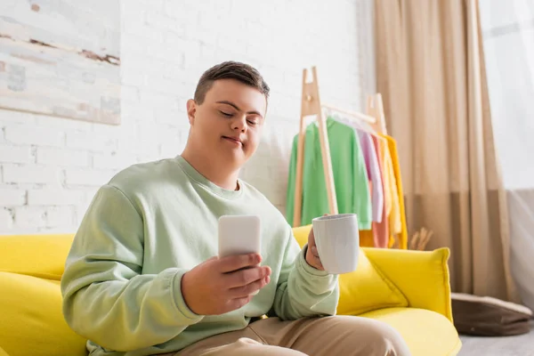Ragazzo adolescente con sindrome di Down utilizzando smartphone e tenendo la tazza sul divano a casa — Foto stock