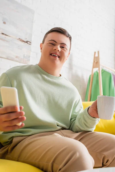 Low angle view of cheerful teenager with down syndrome holding cellphone and cup on couch — Stock Photo