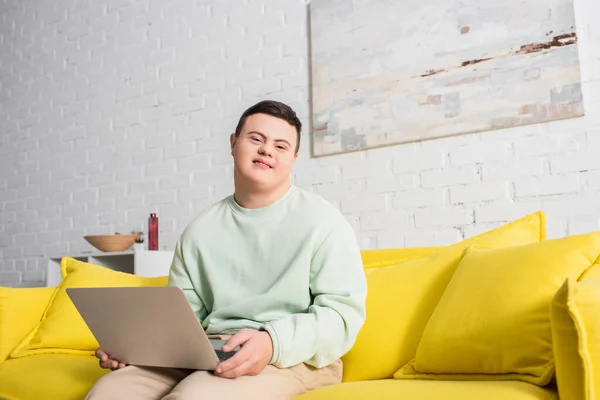 Lächelnder Teenager mit Down-Syndrom hält Laptop in der Hand und blickt auf Couch in die Kamera — Stockfoto