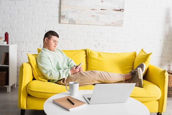 Adolescente con síndrome de Down usando teléfono inteligente en el sofá cerca de la computadora portátil y la taza en la mesa de café en casa - foto de stock