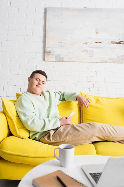 Teenager mit Down-Syndrom hält Handy in der Nähe verschwommener Tasse und Laptop zu Hause — Stockfoto