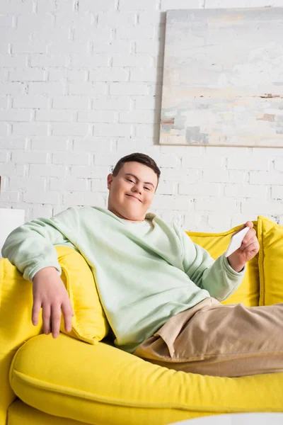 Positive teenager with down syndrome holding smartphone while sitting on couch at home — Stock Photo