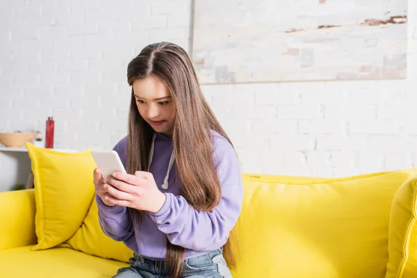 Chica adolescente con síndrome de Down usando teléfono inteligente en el sofá en casa - foto de stock