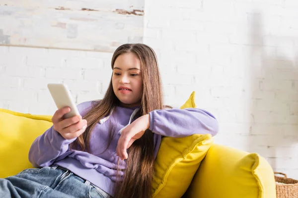 Adolescente sorridente com síndrome semeada usando telefone celular desfocado no sofá em casa — Fotografia de Stock