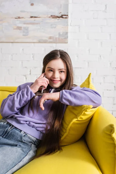 Positiva chica adolescente con síndrome de Down hablando en el teléfono inteligente y mirando a la cámara en casa - foto de stock