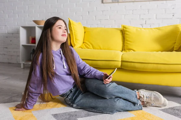 Teen girl with down syndrome holding smartphone while sitting on floor at home — Stock Photo