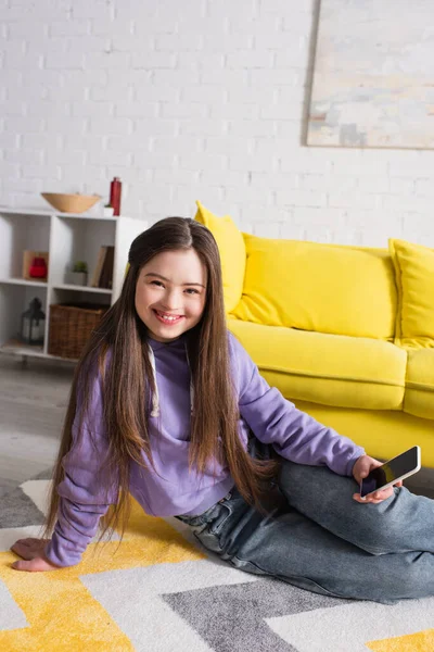 Cheerful girl with down syndrome holding cellphone while sitting on floor at home — Stock Photo