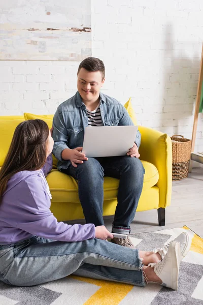 Teenie-Mädchen mit Down-Syndrom hält Smartphone in der Hand und spricht mit Freund mit Laptop auf Couch zu Hause — Stockfoto