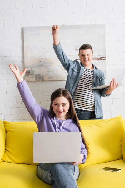 Adolescentes emocionados con síndrome de Down sosteniendo computadoras portátiles en casa - foto de stock