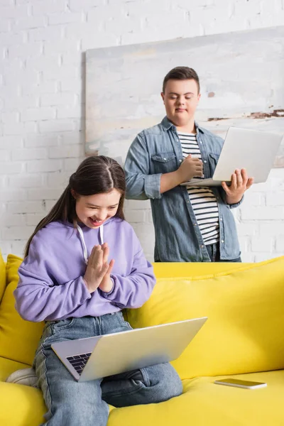 Cheerful teenager with down syndrome using laptop near smartphone and friend at home — Stock Photo