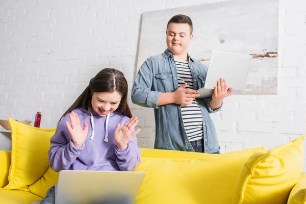 Happy teenager with down syndrome having video call on laptop near blurred friend at home — Stock Photo