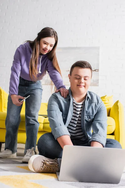 Teenager with down syndrome using laptop near girlfriend with cellphone at home — Stock Photo