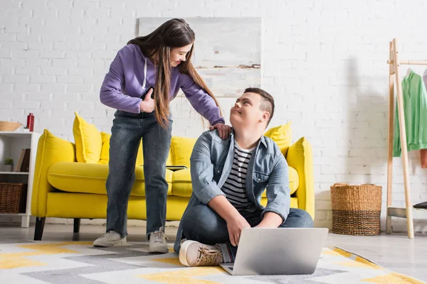 Sonriente chica adolescente con síndrome de Down sosteniendo teléfono inteligente cerca de novio y portátil en el suelo en casa - foto de stock