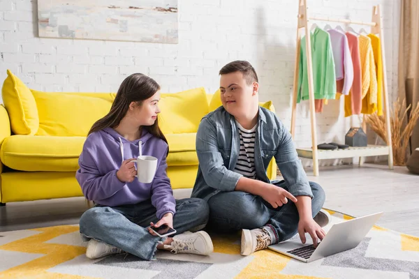 Teen boy with down syndrome pointing at laptop near friend with cup and cellphone at home — Stock Photo