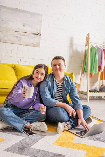 Adolescente sorridente con tazza che guarda la fotocamera vicino al fidanzato e ai dispositivi a casa — Foto stock