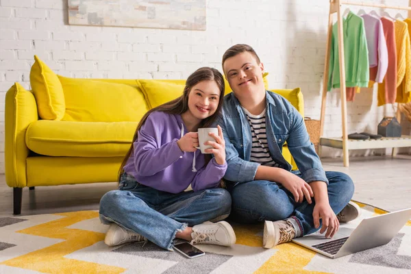 Positive couple of teenagers with down syndrome holding cup and using laptop at home — Stock Photo