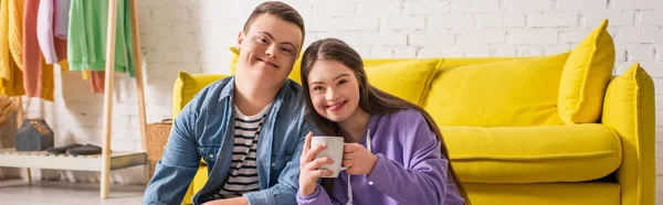 Smiling teen girl holding cup and looking at camera near boyfriend at home, banner — Stock Photo