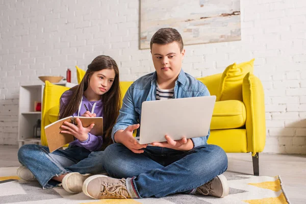 Teenager with down syndrome writing on notebook near friend with laptop at home — Stock Photo