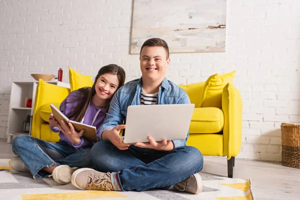 Позитивні підлітки з синдромом Дауна тримають ноутбук і блокнот на підлозі вдома — стокове фото