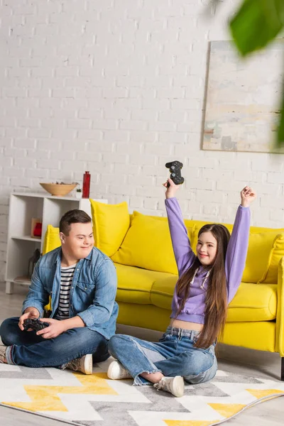 KYIV, UCRANIA - 21 de enero de 2022: Adolescente excitada con síndrome de Down sosteniendo el joystick cerca de un amigo en casa - foto de stock