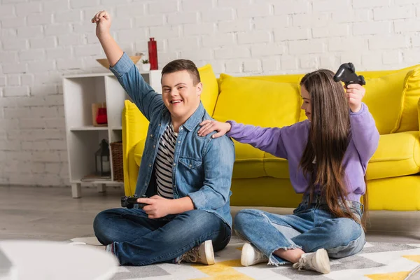 KYIV, UKRAINE - JANUARY 21, 2022: Positive boy with down syndrome showing yes gesture while playing video game near friend at home — Stock Photo