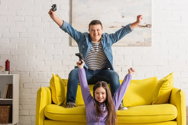KYIV, UKRAINE - JANUARY 21, 2022: Cheerful teenagers with down syndrome showing yes gesture while playing video game at home — Stock Photo