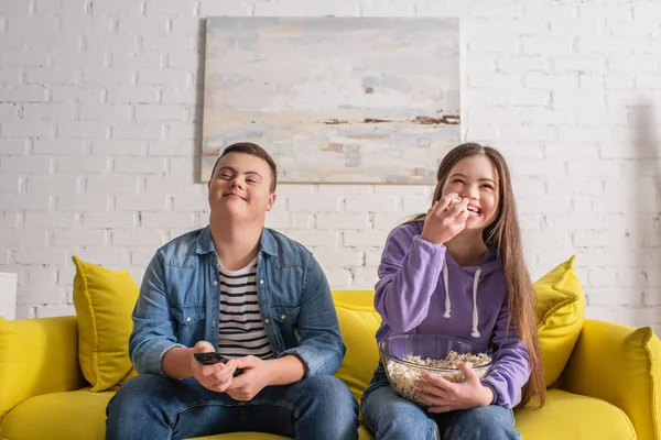 Adolescente positiva con síndrome de Down sosteniendo palomitas de maíz cerca de un amigo con controlador remoto en casa - foto de stock