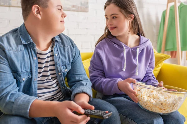 Улыбающиеся подростки с синдромом Дауна держат дома попкорн и пульт дистанционного управления — стоковое фото
