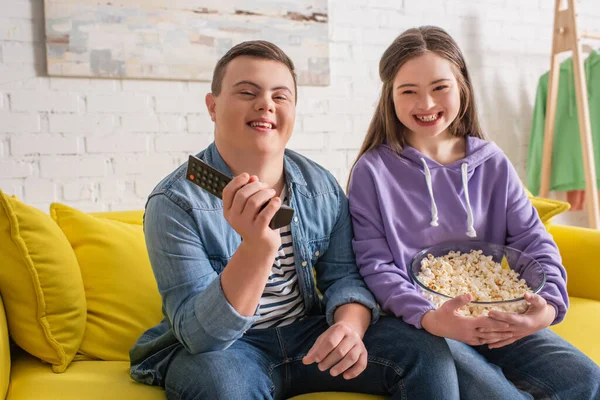 Happy teenagers with down syndrome holding remote controller and popcorn on couch — Stock Photo